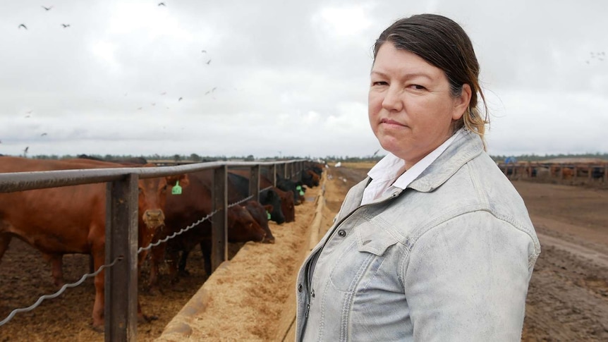 A woman stands in front of a cattle feedlot and looks at the camera, cows eat in the background.