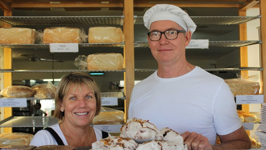 A woman and a man stand behind a counter stacked with bakery items eg bread, meringues, cake