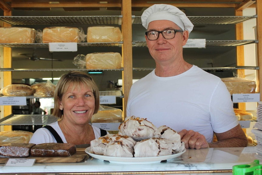 A woman and a man stand behind a counter stacked with bakery items eg bread, meringues, cake