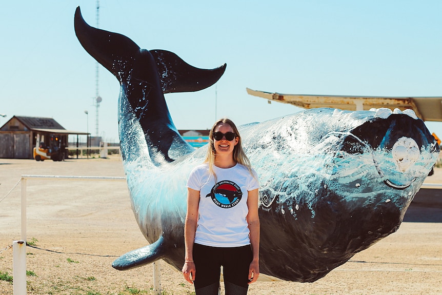 A smiling young woman stands at a remote roadhouse in front of a model of a whale.