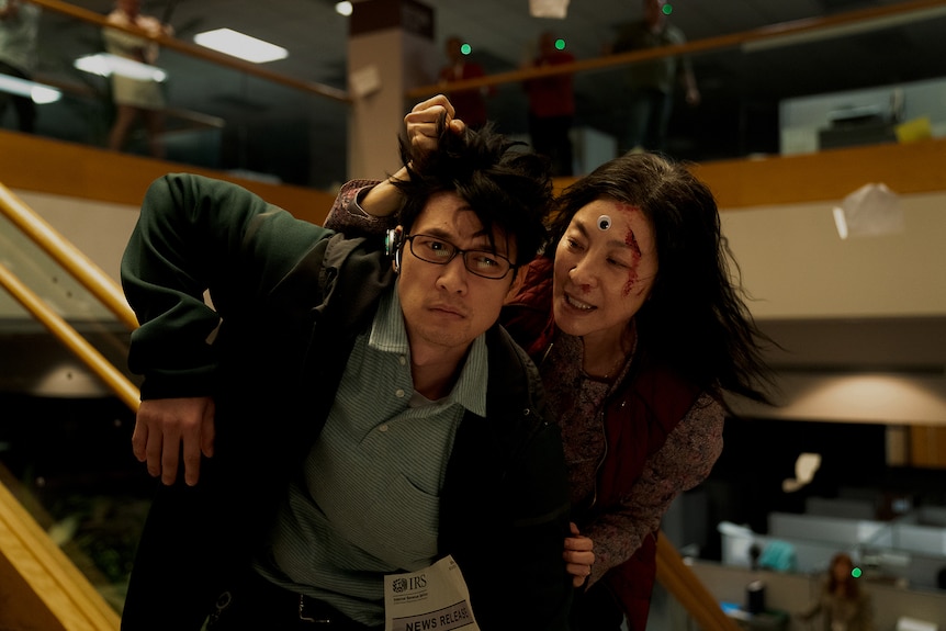 Chinese-American woman with a plastic eye and blood on her forehead grabs hair of Asian man wearing headphones and glasses.