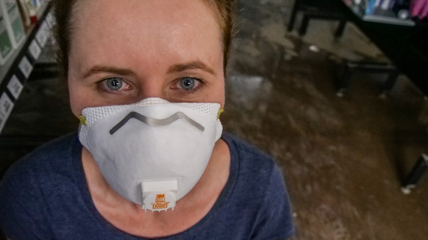 A woman wearing a face mask looking directly into the camera. Behind her is a water-covered floor.