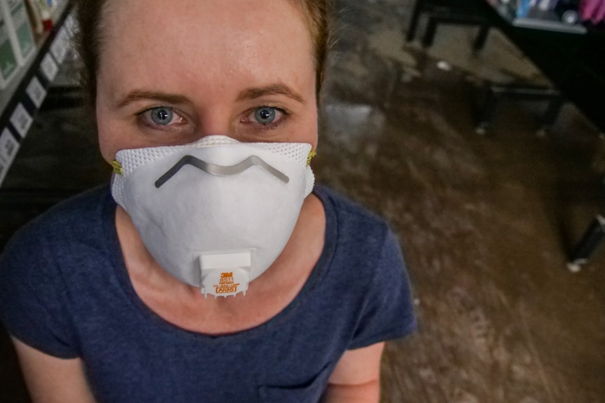 A woman wearing a face mask looking directly into the camera. Behind her is a water-covered floor.