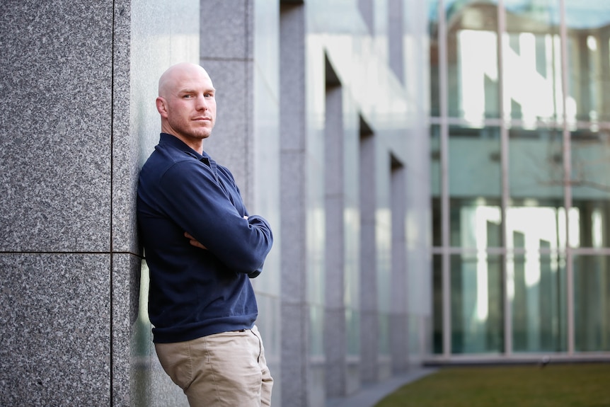 David Pocock wears a navy blue sweater while leaning on the wall of the Parliament House courtyard