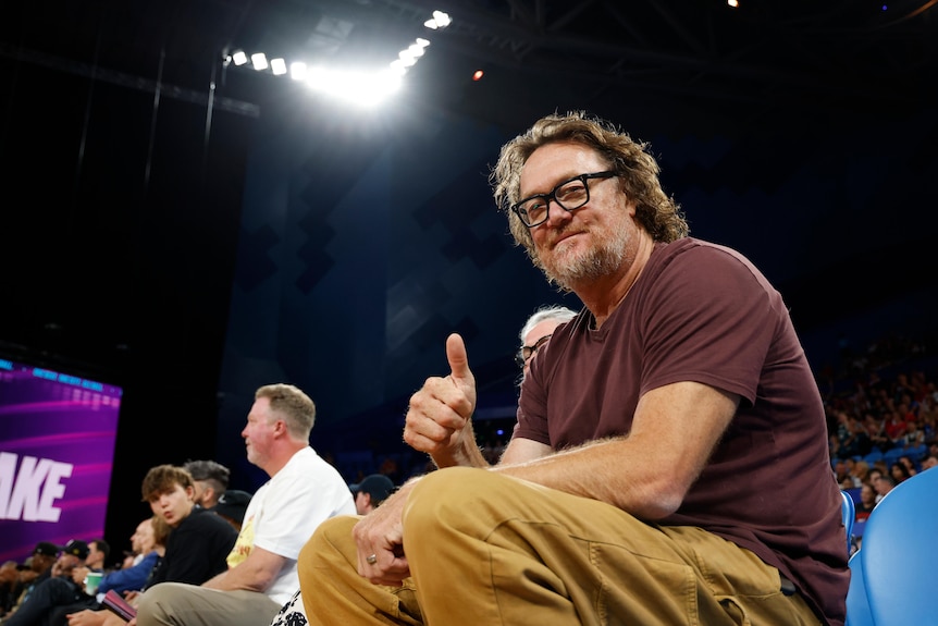 Former basketball player sitting courtside, giving a thumbs-up to the camera, smiling