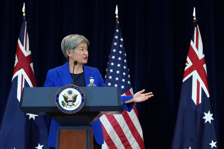 A woman gestures towards a man with whom she stands upon a stage before Australian and US flags.