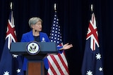A woman gestures towards a man with whom she stands upon a stage before Australian and US flags.