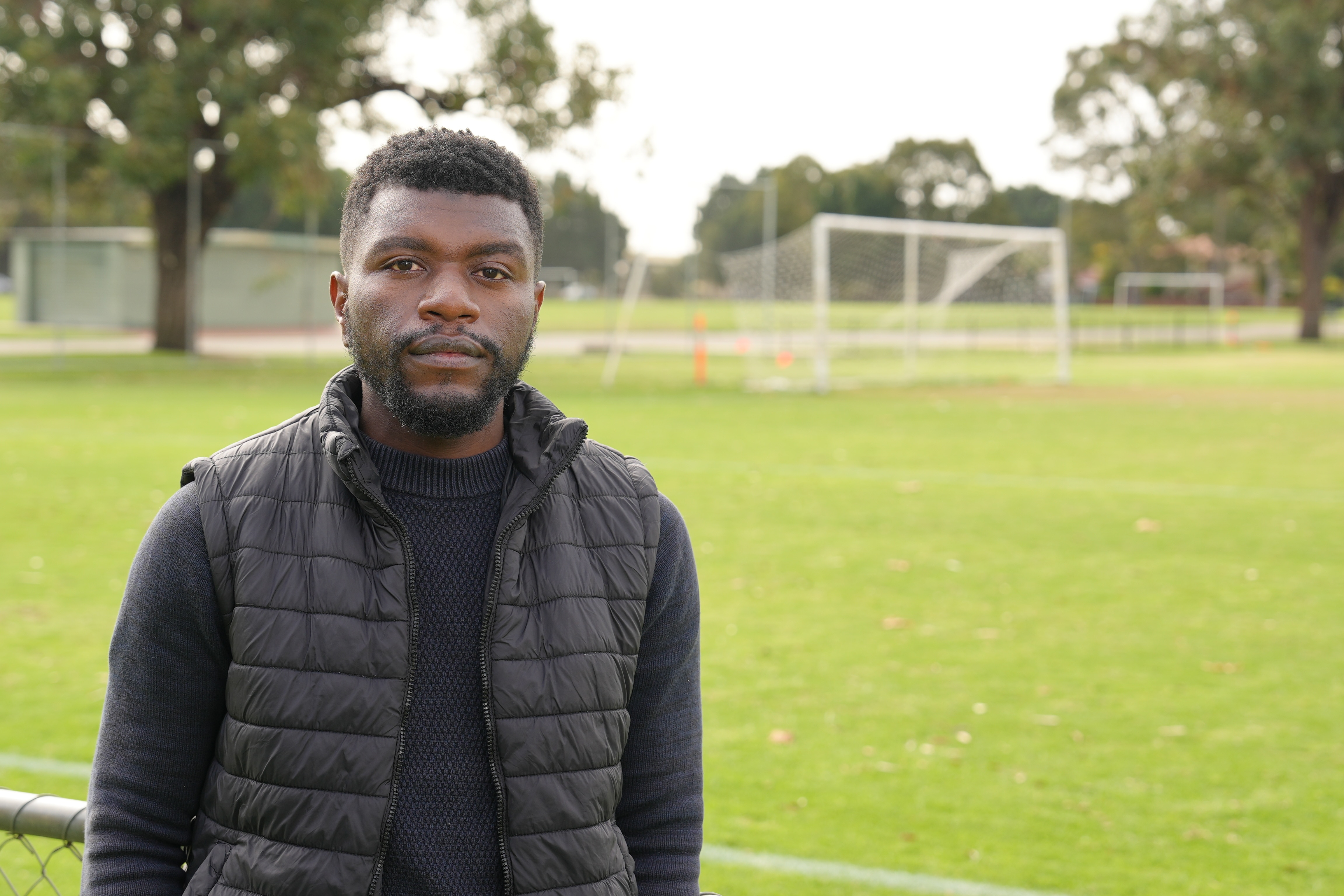 A man wears black clothing and stands in front of a soccer goal on a field.