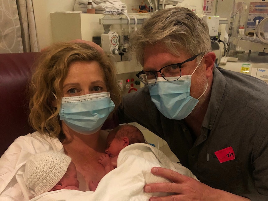 A masked woman and man in hospital, two premature babies are on the woman's chest in a blanket.