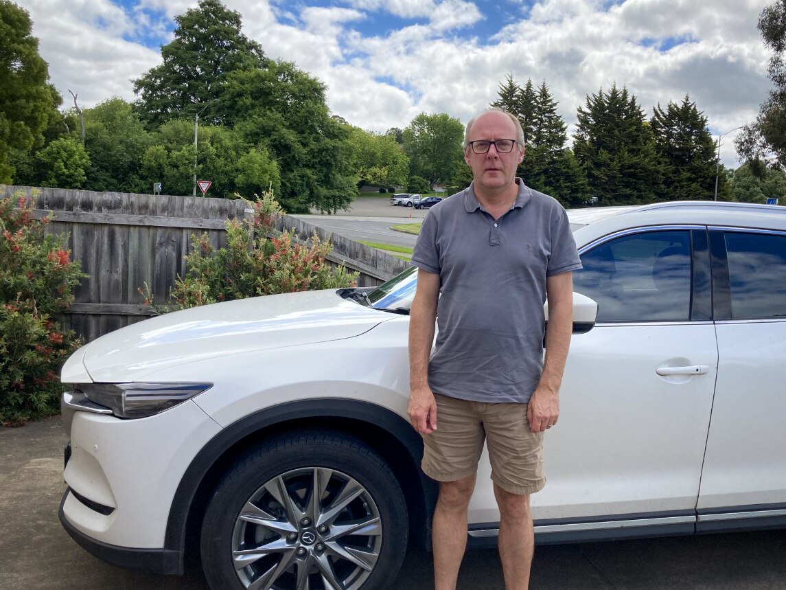 An older man with white hair and black glasses stands in front of a white car.