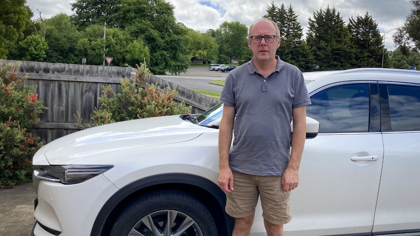 An older man with white hair and black glasses stands in front of a white car.