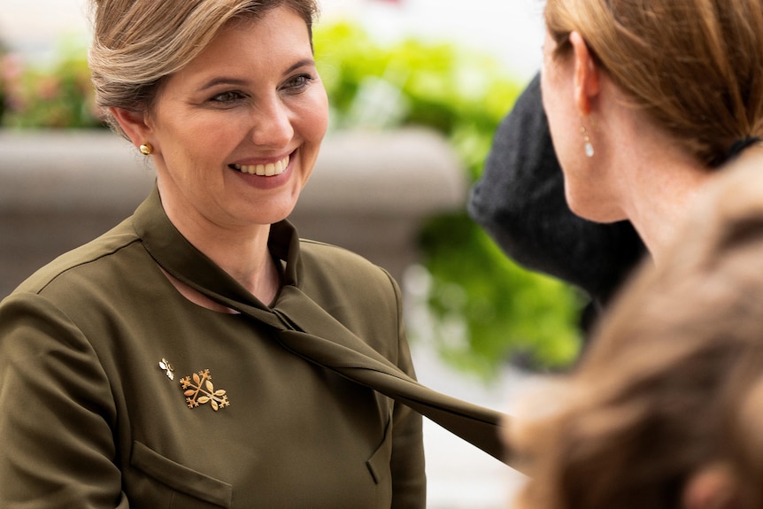 A blonde woman with a green dress smiles at another woman. There are military badges on her dress.