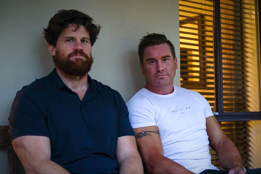 Two men sit on a bench in front of window blinds