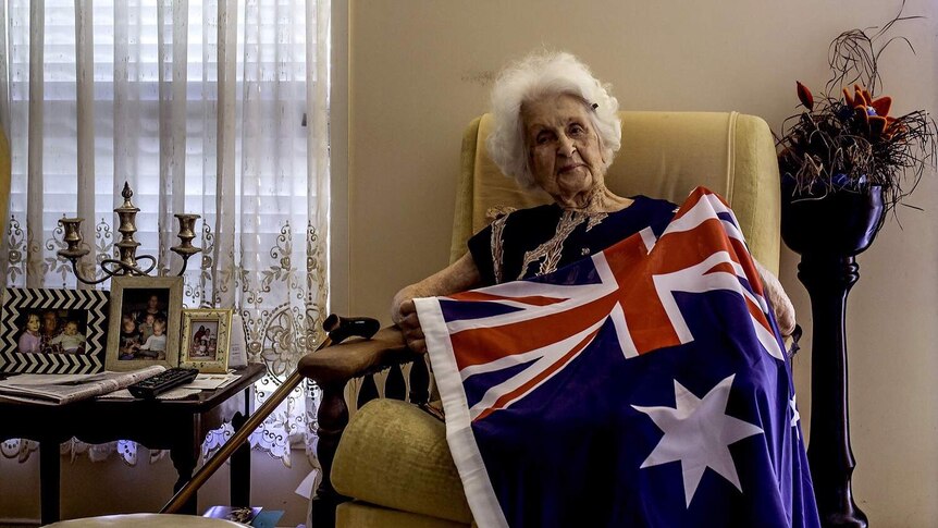 An elderly woman sits on a chair, the Australian Flag draped across her.