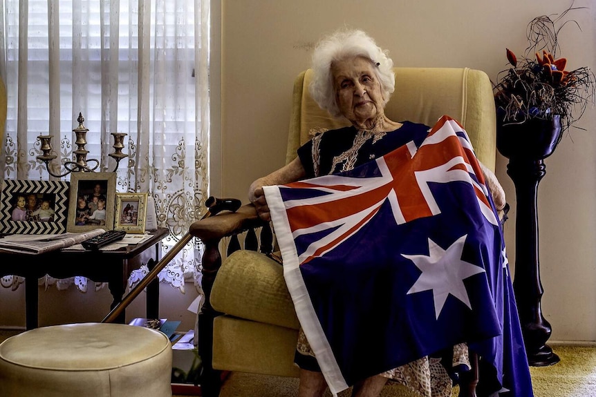 An elderly woman sits on a chair, the Australian Flag draped across her.