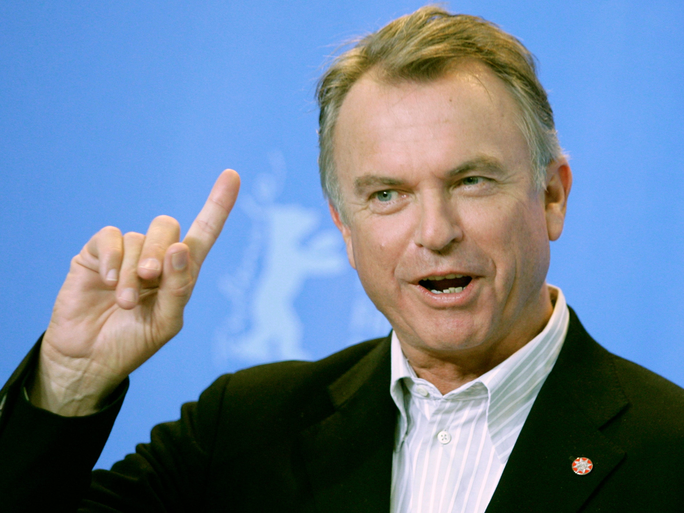 Sam Neill mid-speech with his right index finger raised, in front of a blue backdrop while wearing a black blazer.