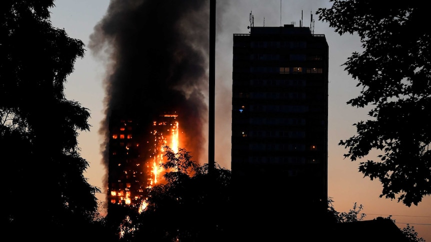 A dusk shot shows a burning building tower in the background as another high rise is in shadows in the foreground.