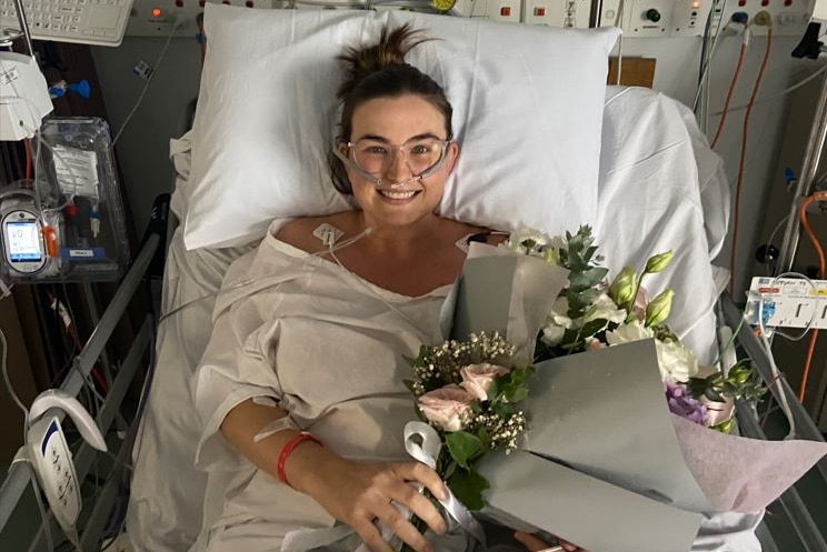 Woman lays in hospital bed, smiling and holding a bouquet of flowers