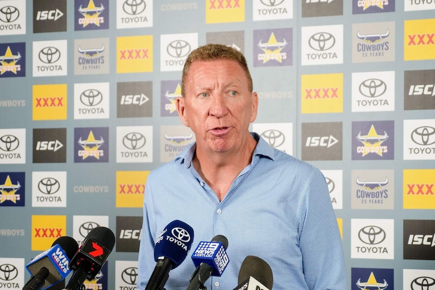 A middle aged man standing in front of microphones and a North Queensland Cowboys banner.