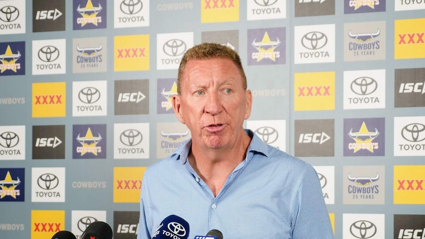 A middle aged man standing in front of microphones and a North Queensland Cowboys banner.