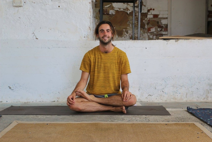 A man sits smiling with cross legs. He is on a yoga mat.