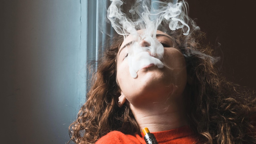 A young woman with dark curly hair, orange top, holds an e-cigarette, leans back and expels smoke.