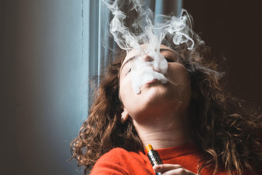 A young woman with dark curly hair, orange top, holds an e-cigarette, leans back and expels smoke.