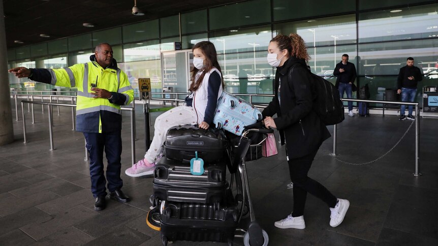 A man in high-visibility workwear directs a masked woman and her daughter, sitting on a trolley full of bags, at the airport.