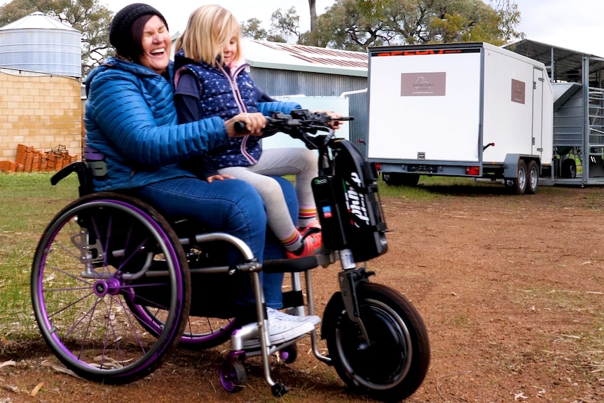 Woman in wheelchair laughing with child on lap and farm setting in background.