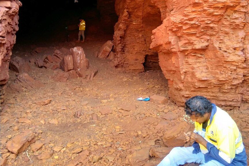 An Aboriginal man in jeans and a hi-vis shirt sits at the entrance to a cave with another man standing in the mouth of the cave.