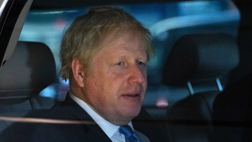 Boris Johnson looks straight ahead as he sits in the back of a car. He has a worried expression on his face and wears a suit.
