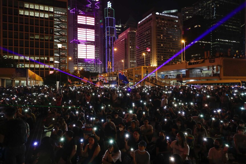 A large crowd of people sit in a city square holding up lit-up mobile phones like candles at a prayer service.