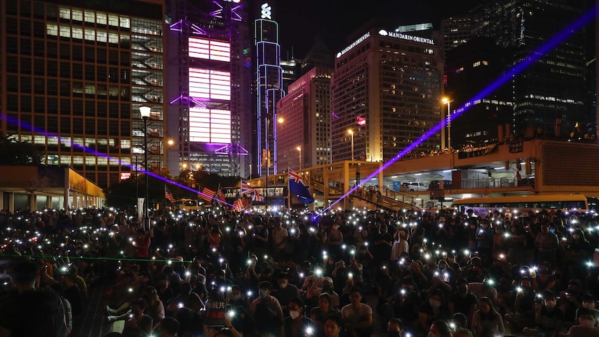 A large crowd of people sit in a city square holding up lit-up mobile phones like candles at a prayer service.