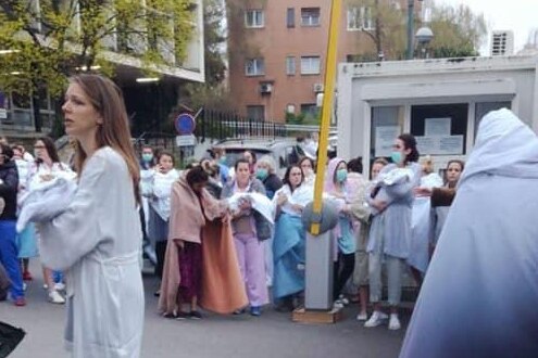 You view a crowd of mothers in hospital gowns and surgical masks carrying newborns in blankets outside a hospital building.