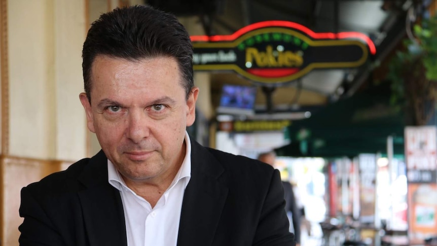 Nick Xenophon stands outdoors with a pokies sign in the background.