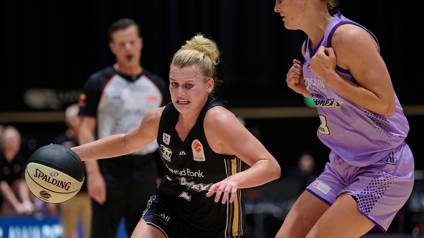 A WNBL basketballer grimaces as she runs downcourt with the ball trying to go around a defender during a game.