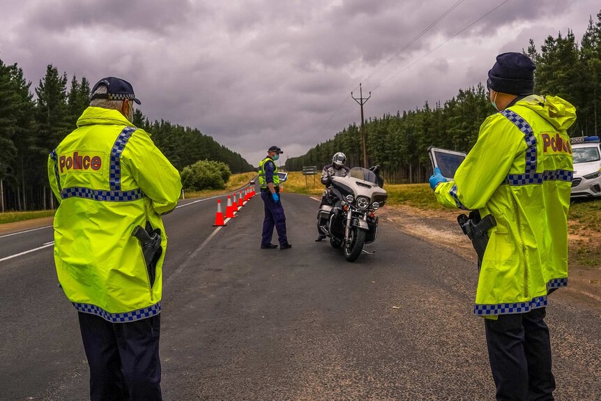 Two police in yellow jackets stand in the foreground and another is farther back with a police officer on a motorcycle