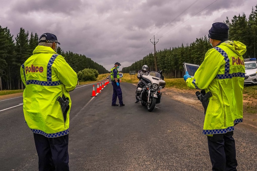 Two police in yellow jackets stand in the foreground and another is farther back with a police officer on a motorcycle