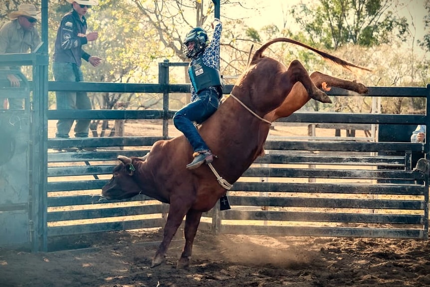A young bull riding with one hand in the air and holding onto a big brown bull bucking on red dirt.