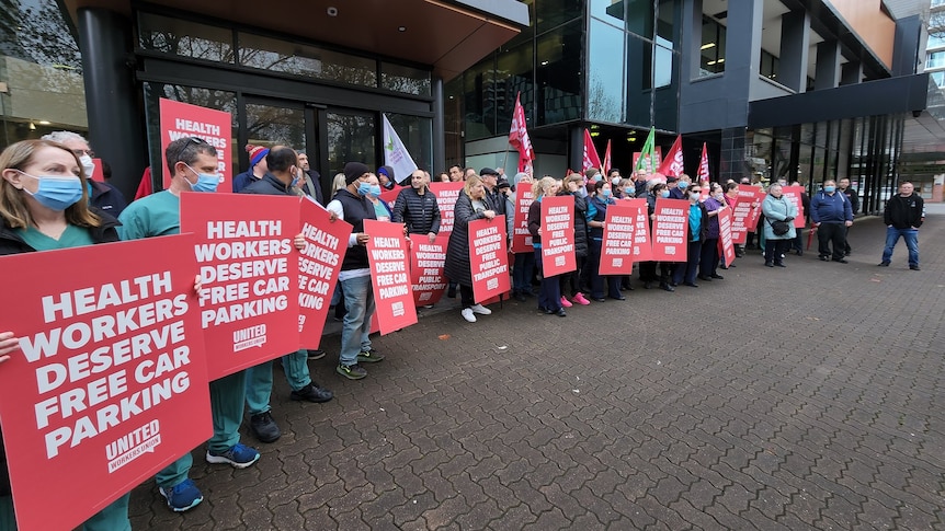 Protestors holding red placards saying 'health workers deserve free car parking' on a footpath