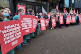 Protestors holding red placards saying 'health workers deserve free car parking' on a footpath