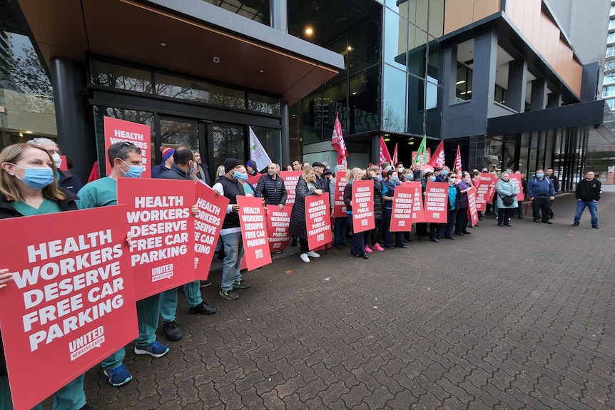 Protestors holding red placards saying 'health workers deserve free car parking' on a footpath