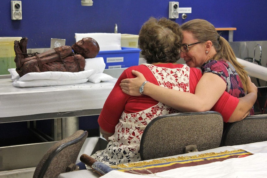 Two women embrace after a mummified body is presented inside a laboratory.