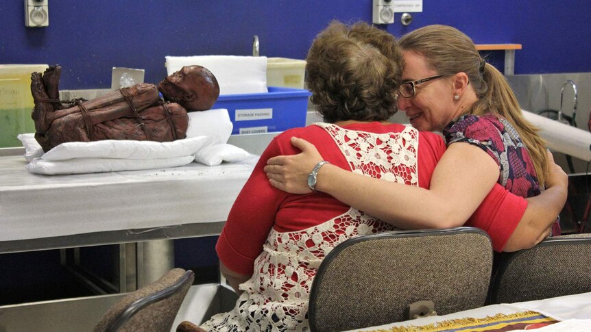 Two women embrace after a mummified body is presented inside a laboratory.