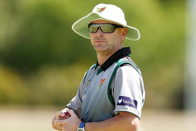 A man wearing sunglasses and a white wide-brimmed hat holds a cricket ball with both hands. 