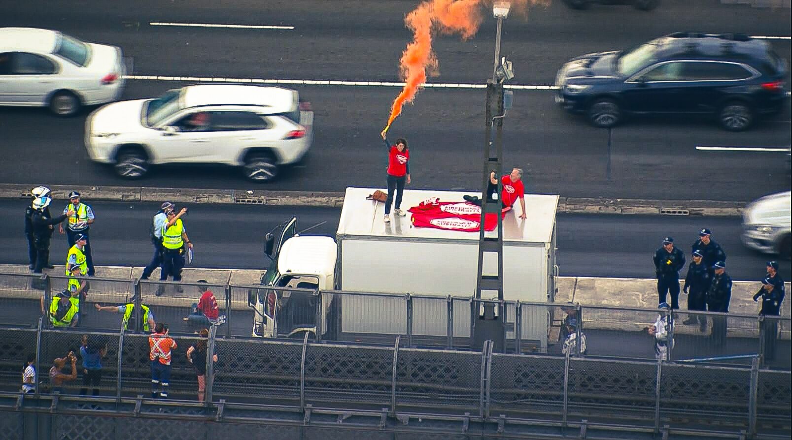 Two people wearing red on top of a truck letting off a flare, in the middle of traffic with police surrounding them.