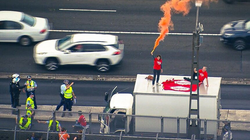 Two people wearing red on top of a truck letting off a flare, in the middle of traffic with police surrounding them.
