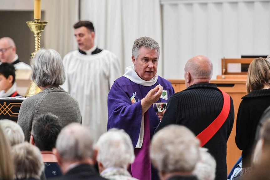 Dean Lawrence Kimberley wears purple robes as he stands opposite a churchgoer with a goblet in his hand.