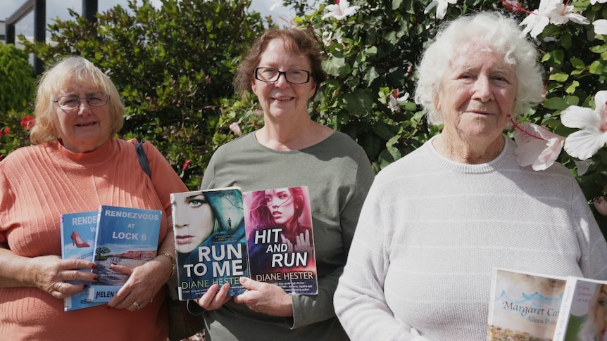 Three women holding books they have written in front of a tree with flowers.