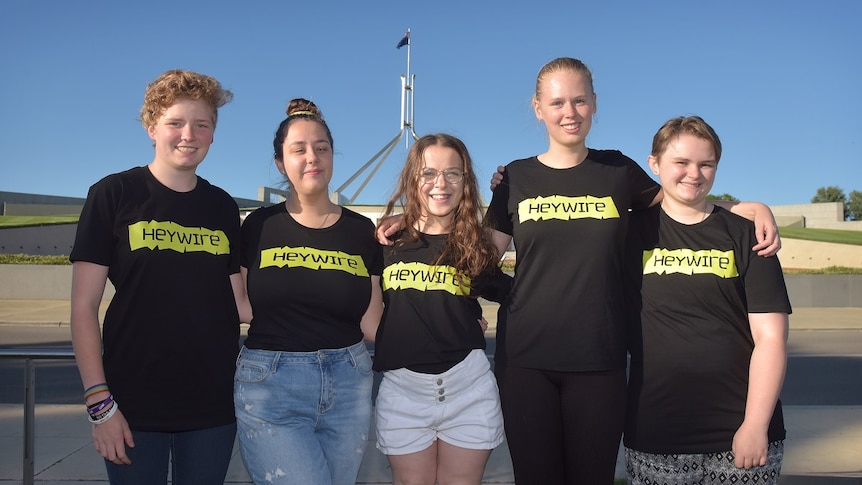 Five young women in Heywire shirts stand in front of Parliament House with arms around each other.